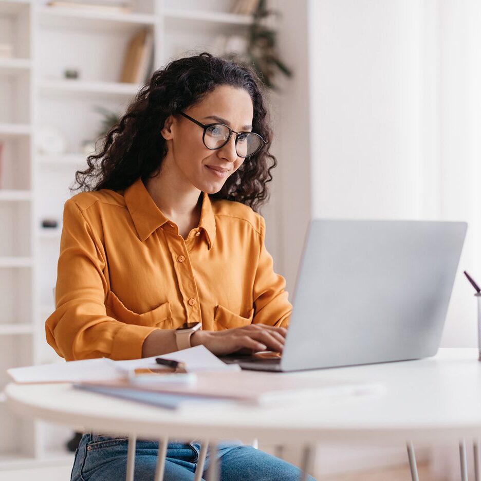 A person wearing glasses and a yellow shirt is sitting at a table, typing on a laptop in a bright room with shelves in the background.